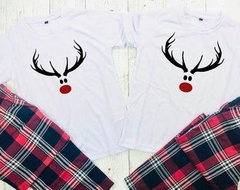 May include: Two white t-shirts featuring a black reindeer antler design, black eyes, and a red nose, alongside red, black, and white plaid pajama bottoms. A festive matching pajama set for Christmas.