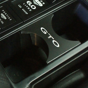 May include: Black cup holder with a silver GTO logo. The cup holder is in a car's center console. The console has a black background with silver buttons and text that reads "T/C", "PONTIAC GTO", "FUEL FOR THE SOUL", "6.0 LITRE", and "LS2 400HP".