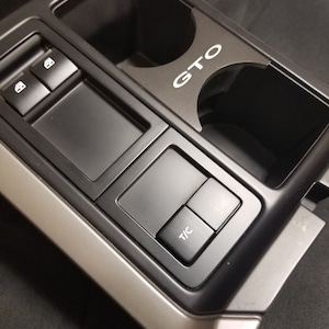 May include: Black plastic car interior trim panel with a cup holder and a button labeled "T/C". The panel also has a silver trim and the word "GTO" printed on it.