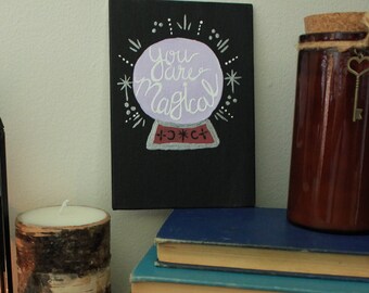 May include: A black metal lantern with a glass panel and a candle inside. There are books stacked on a shelf with a brown glass jar with a key attached to it. A sign with the text "You are Magical" is on the wall behind the books.