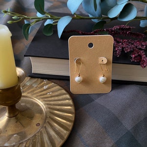 May include: A pair of gold earrings with white pearl drops on a beige card. The card is on a brown and grey plaid surface.