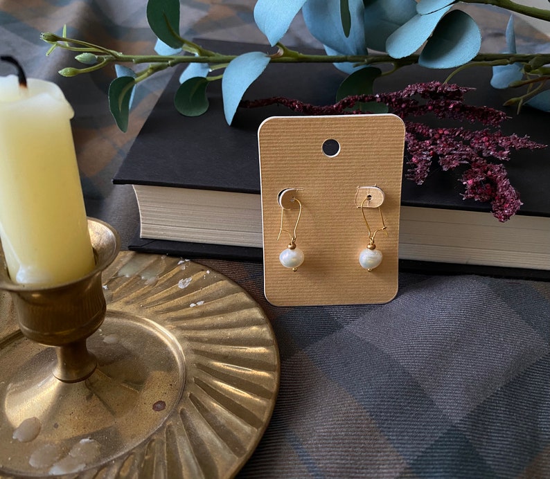 May include: A pair of gold earrings with white pearl drops on a beige card. The card is on a brown and grey plaid surface.