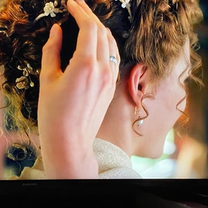 May include: A woman with curly brown hair is having her hair styled. She is wearing a white dress and has a pearl earring in her left ear. Her hair is decorated with a floral hairpiece.