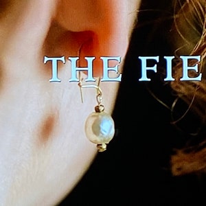 May include: A close-up of a person's ear with a gold earring featuring a white pearl. The text "THE FIE" is visible in the background.
