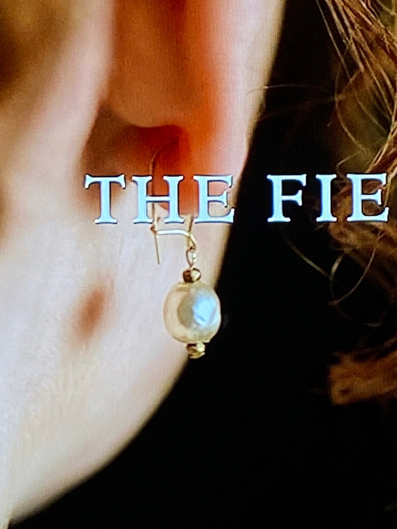 May include: A close-up of a person's ear with a gold earring featuring a white pearl. The text "THE FIE" is visible in the background.