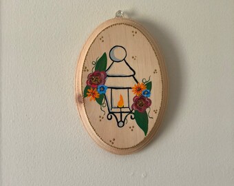 May include: A wooden oval sign featuring a painted image of a lantern with a flame inside. The lantern is surrounded by flowers in shades of red, orange, blue, and yellow.