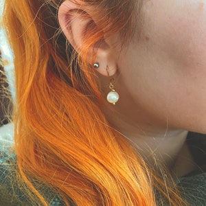 May include: A close-up of a person's ear with a gold earring featuring a single white pearl. The earring is dangling from the earlobe.