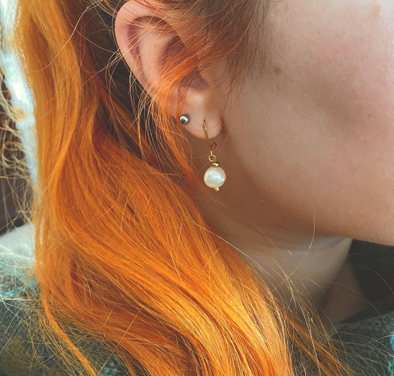 May include: A close-up of a person's ear with a gold earring featuring a single white pearl. The earring is dangling from the earlobe.