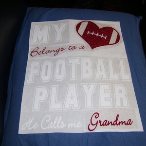 May include: White vinyl decal with the text "My Belongs to a Football Player He Calls me Grandma" in a glittery white font. A red glitter heart with a white football graphic is in the upper right corner.