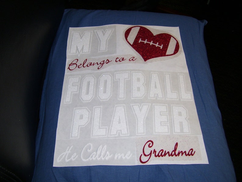 May include: White vinyl decal with the text "My Belongs to a Football Player He Calls me Grandma" in a glittery white font. A red glitter heart with a white football graphic is in the upper right corner.