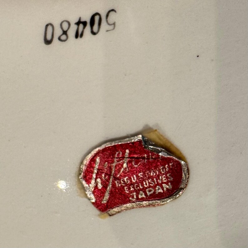 May include: Close-up of a white ceramic surface with the number "50409" printed in black. A red and silver sticker with the text "REG. U.S. PAT. OFF. EXCLUSIVES JAPAN" is affixed to the surface.