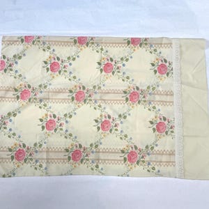 May include: Cream-colored pillowcase with a floral pattern featuring pink roses, green leaves, and small blue and yellow flowers. The design is set against a diamond grid with brown stripes. A white lace trim adorns one edge.