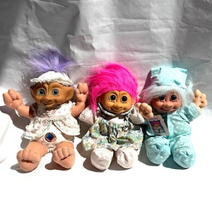 May include: Three troll dolls with tan skin and large eyes. The left doll has purple hair and a white floral outfit with a blue gem. The center doll has bright pink hair and a floral dress. The right doll has light blue hair and a light blue polka dot pajama set.