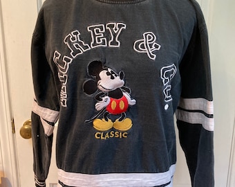 May include: Dark grey sweatshirt with white and black striped trim. Features an embroidered Mickey Mouse and the text "GREY & Co. CLASSIC". The long-sleeved shirt has white stripes on the sleeves and bottom.
