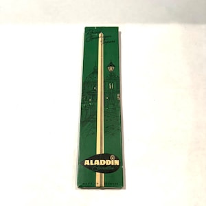 May include: A green box of Aladdin Candles with a gold and cream-colored candle inside. The box features a drawing of a building with a dome and a street lamp. The text "ALADDIN Candles" is printed on the box, along with "MADE IN DENMARK".