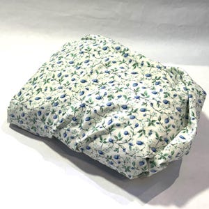 May include: A white fabric item with a repeating pattern of blue berries and green leaves on vines. The fabric is slightly crumpled, suggesting it is a pillowcase or similar item.
