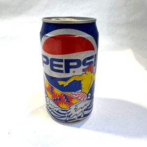 May include: Vintage Pepsi soda can with a blue, red, and white design. The can features the Pepsi logo and a graphic of a surfer riding a wave. The can is cylindrical and made of metal.