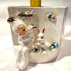 May include: A white ceramic planter with a whimsical design. It features a seated figure with a gold-tipped hat and a small white rabbit. Pink and blue floral accents adorn the planter's surface.