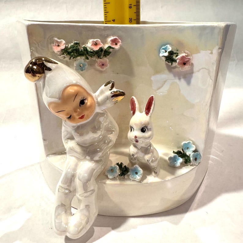 May include: A white ceramic planter with a whimsical design. It features a seated figure with a gold-tipped hat and a small white rabbit. Pink and blue floral accents adorn the planter's surface.