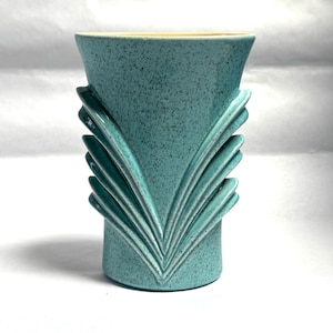 May include: A teal ceramic vase with a speckled finish. The vase features a flared top and a stylized, symmetrical design of layered, curved elements on the sides. The interior rim is a light yellow color.