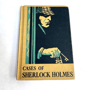 May include: A hardcover book titled Cases of Sherlock Holmes with a yellow spine and cover. The front cover features a dark blue graphic illustration of a person wearing a plaid deerstalker hat, holding a set of keys in a yellow and teal color scheme.