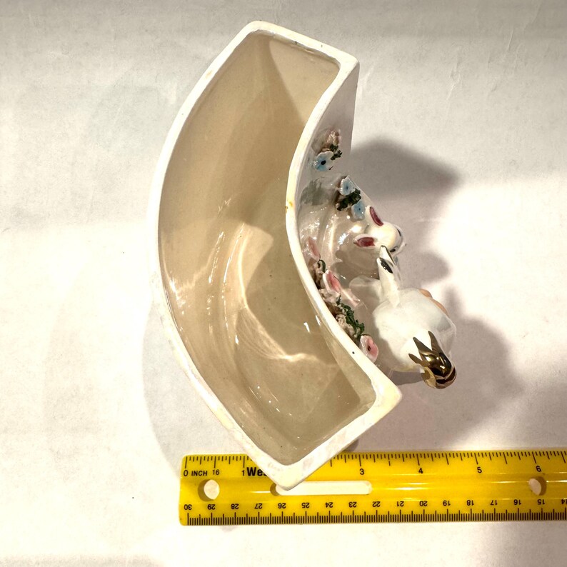 May include: A curved, off-white ceramic vase with a decorative swan and floral arrangement. The swan has gold accents. The vase is approximately 6 inches long. The flowers are pink, blue, and green.