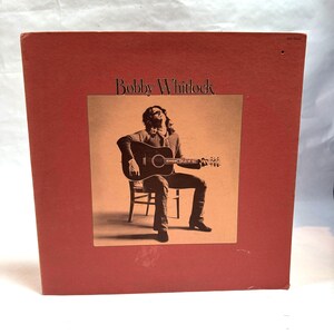 May include: A red vinyl record album cover with the title "Bobby Whitlock" in white text. The cover features a photo of a man sitting on a chair, playing a brown acoustic guitar.
