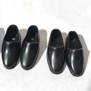 May include: Four pairs of black plastic doll shoes. The shoes are all the same style, with a rounded toe and a simple design.