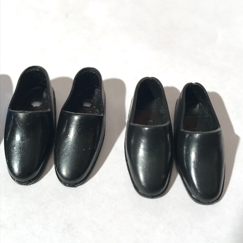 May include: Four pairs of black plastic doll shoes. The shoes are all the same style, with a rounded toe and a simple design.