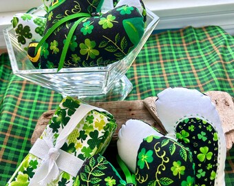 May include: Heart-shaped St. Patrick's Day decorations in a glass bowl and on a wooden surface. The hearts are black with green shamrocks and a green ribbon. Other decorations include green and white shamrock-patterned fabric.