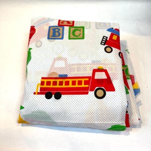 May include: Folded white fabric with a pattern of red fire trucks, ABC blocks, and other colorful shapes. The fabric has a dotted background. The fire trucks have yellow ladders and black wheels.