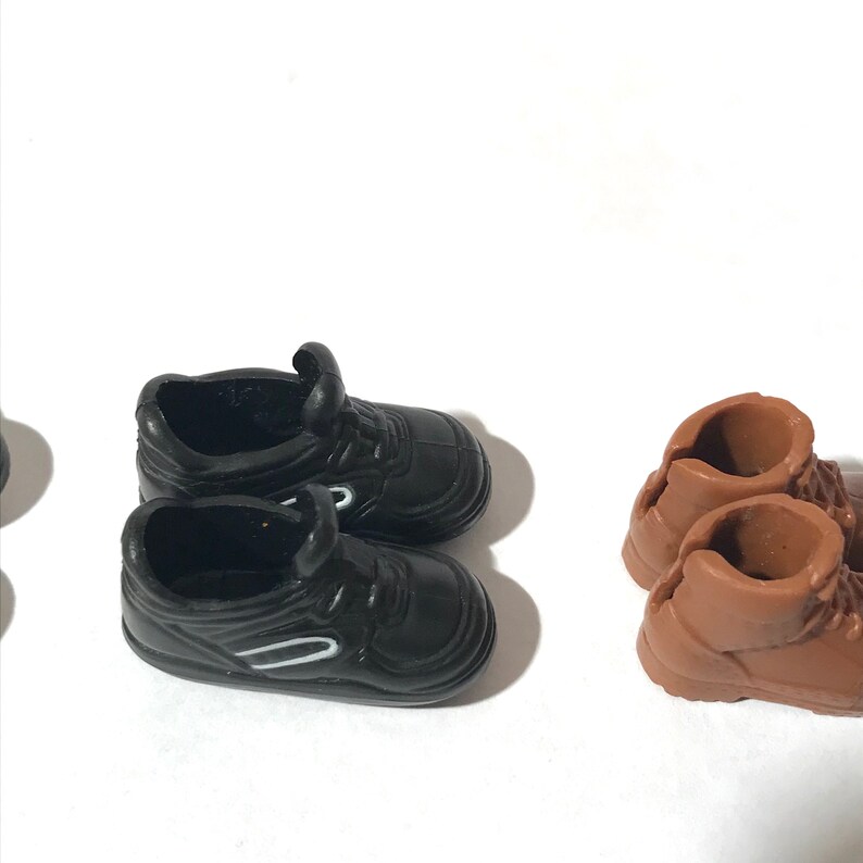 Op de afbeelding: Twee paar poppen schoenen. Een paar is zwarte sneakers met witte accenten. Het andere paar is bruine wandelschoenen.