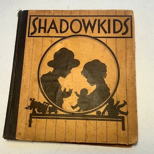 May include: A vintage book titled "Shadow Kids" with a brown cover and a black spine. The cover features a silhouette of two children and a baby in a circle. The book has a striped pattern on the cover.