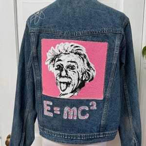 May include: A blue denim jacket with a pink patch featuring a cartoon image of Albert Einstein with his tongue sticking out. The patch also includes the text "E=MC^2" in white with pink glitter.