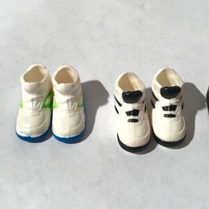 Op de afbeelding: Twee paar witte sneakers met verschillende gekleurde accenten. Het ene paar heeft groene accenten en blauwe afwerking, het andere heeft zwarte accenten en zwarte afwerking.
