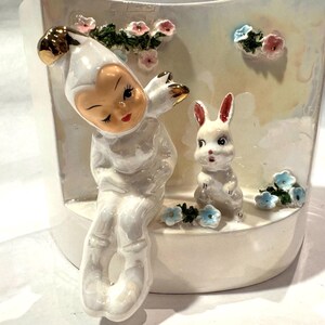 May include: A ceramic planter featuring a whimsical design. It depicts a seated figure in a white outfit with gold accents, winking, alongside a white bunny with red-tipped ears. Pink and blue floral accents decorate the planter, creating a charming, decorative piece.