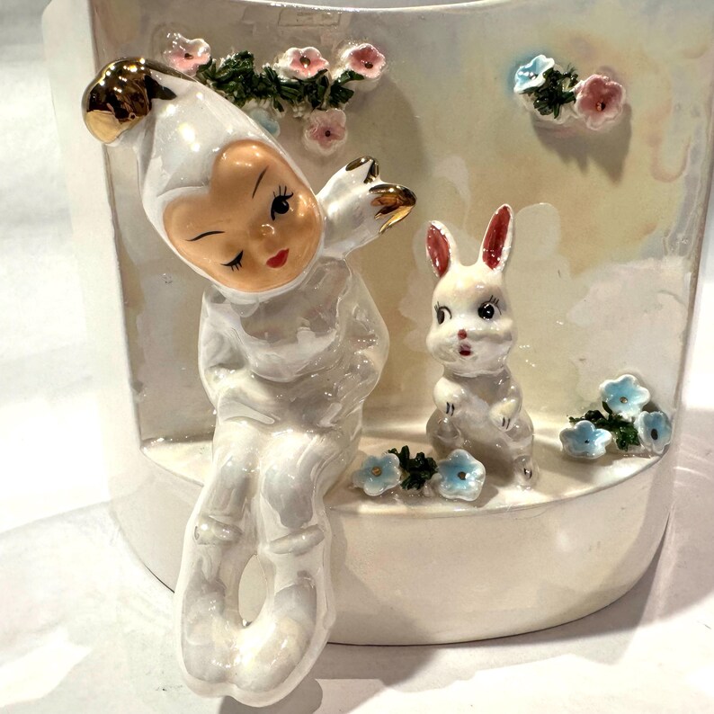 May include: A ceramic planter featuring a whimsical design. It depicts a seated figure in a white outfit with gold accents, winking, alongside a white bunny with red-tipped ears. Pink and blue floral accents decorate the planter, creating a charming, decorative piece.