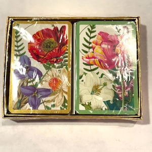May include: Two decks of playing cards in a gold-colored box. Each card deck features a floral design with red poppies, purple irises, and white lilies. The cards are wrapped in clear plastic.