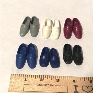May include: Six pairs of miniature plastic doll shoes in various colours, including grey, white, burgundy, blue, and black. The shoes are all loafers with a single strap across the top.