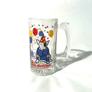 May include: A clear glass beer stein with a colorful design featuring a white bull terrier wearing a party hat and a blue shirt with the word "Spuds" on it. The stein has confetti and balloons on it and the text "Bud Light. Spuds MacKenzie. Original party animal."