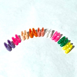 May include: A collection of 12 pairs of colorful plastic doll shoes. The shoes are arranged in a semi-circle, with the toes pointing inwards. The shoes are in various colors, including pink, orange, yellow, green, and white.