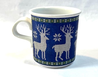 May include: A white ceramic mug with a blue background featuring a pattern of white reindeer and snowflakes. The rim and handle are white. The mug has a green and gold border at the top and bottom of the blue section.