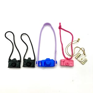 May include: Five toy cameras with straps. Two black cameras with black straps, one blue camera with a purple strap, one pink camera with a pink strap, and one gold camera with a gold strap.