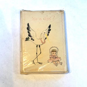May include: A vintage baby announcement card with a white stork carrying a pink baby doll. The card reads "It's a Girl!"