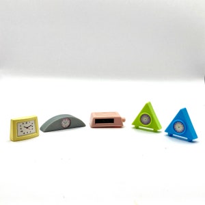 May include: Five miniature clocks in different shapes and colors. A yellow square clock, a gray arched clock, a pink rectangular clock, a green triangular clock, and a blue triangular clock.