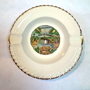 May include: Cream-colored ashtray with a gold-colored, scalloped rim. The center features a colorful illustration of Rock City Gardens, Lookout Mountain. The ashtray has three small indentations for resting cigarettes.