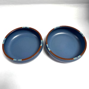 May include: Two blue ceramic bowls with a brown rim and a decorative band of blue and white stripes. The bowls have a smooth, matte finish and are set against a white background. These bowls are suitable for serving food.