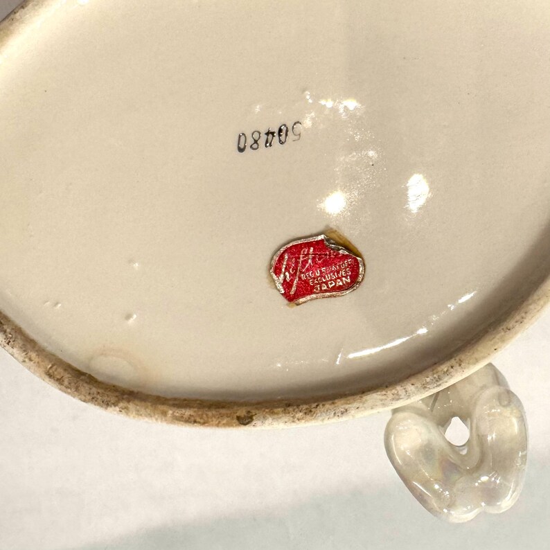 May include: Off-white ceramic piece with a handle. The bottom of the piece has the number "50480" printed in black ink. A red and white sticker with the text "Regus of Exclusives Japan" is affixed to the ceramic.