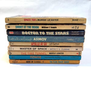 May include: A stack of vintage science fiction paperback books. Titles include "Space Tug," "Shoot at the Moon," "Doctor to the Stars," and "Earth is Room Enough." The books have aged covers in various colors, including yellow, blue, and brown.