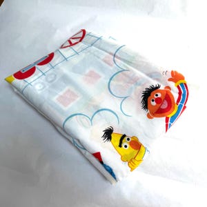 May include: A folded white fabric sheet featuring a repeating pattern of Sesame Street characters Ernie and Bert, alongside red circular shapes and blue grid lines. The print displays the orange-skinned Ernie and yellow-skinned Bert.